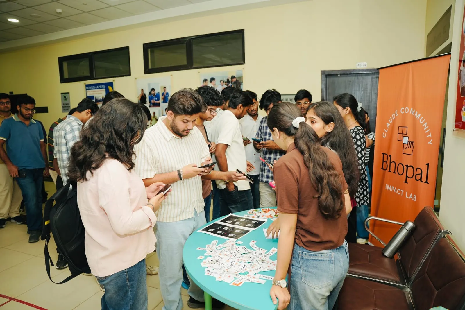 Attendees checking their phones around a table spread with stickers, beside a Claude Community Bhopal Impact Lab banner