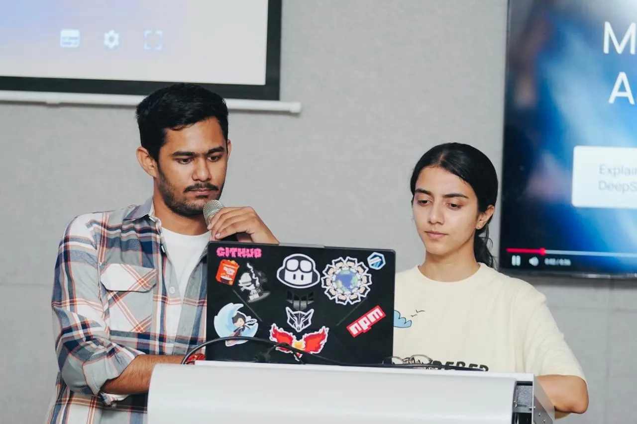 Two presenters at a lectern, one speaking into a microphone behind a sticker-covered laptop