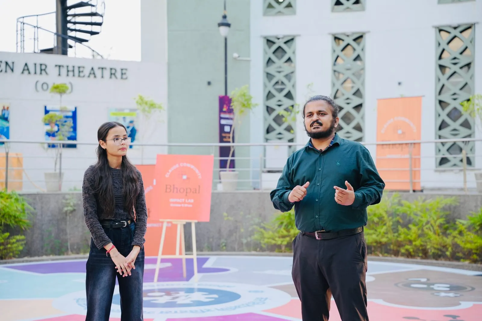 Two presenters in the open-air theatre courtyard, one speaking, beside a Claude Community Bhopal Impact Lab sign