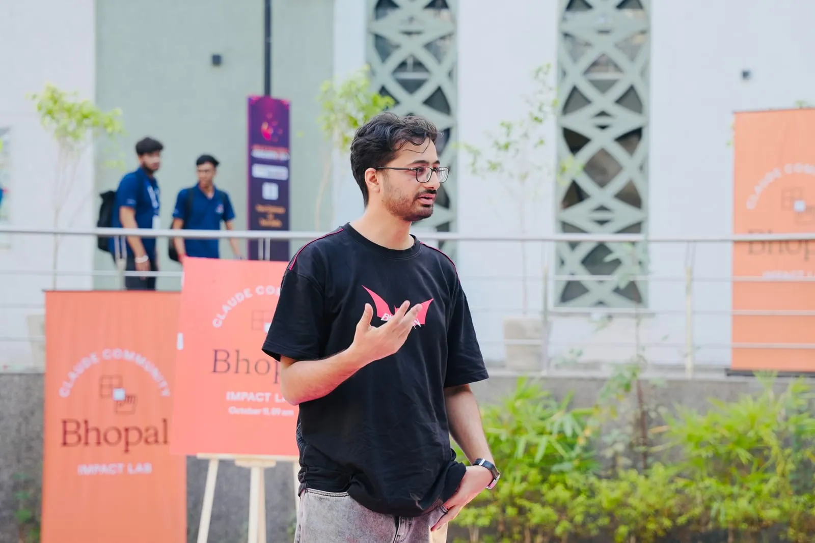 A speaker in a black T-shirt talking outdoors in front of Claude Community Bhopal Impact Lab banners