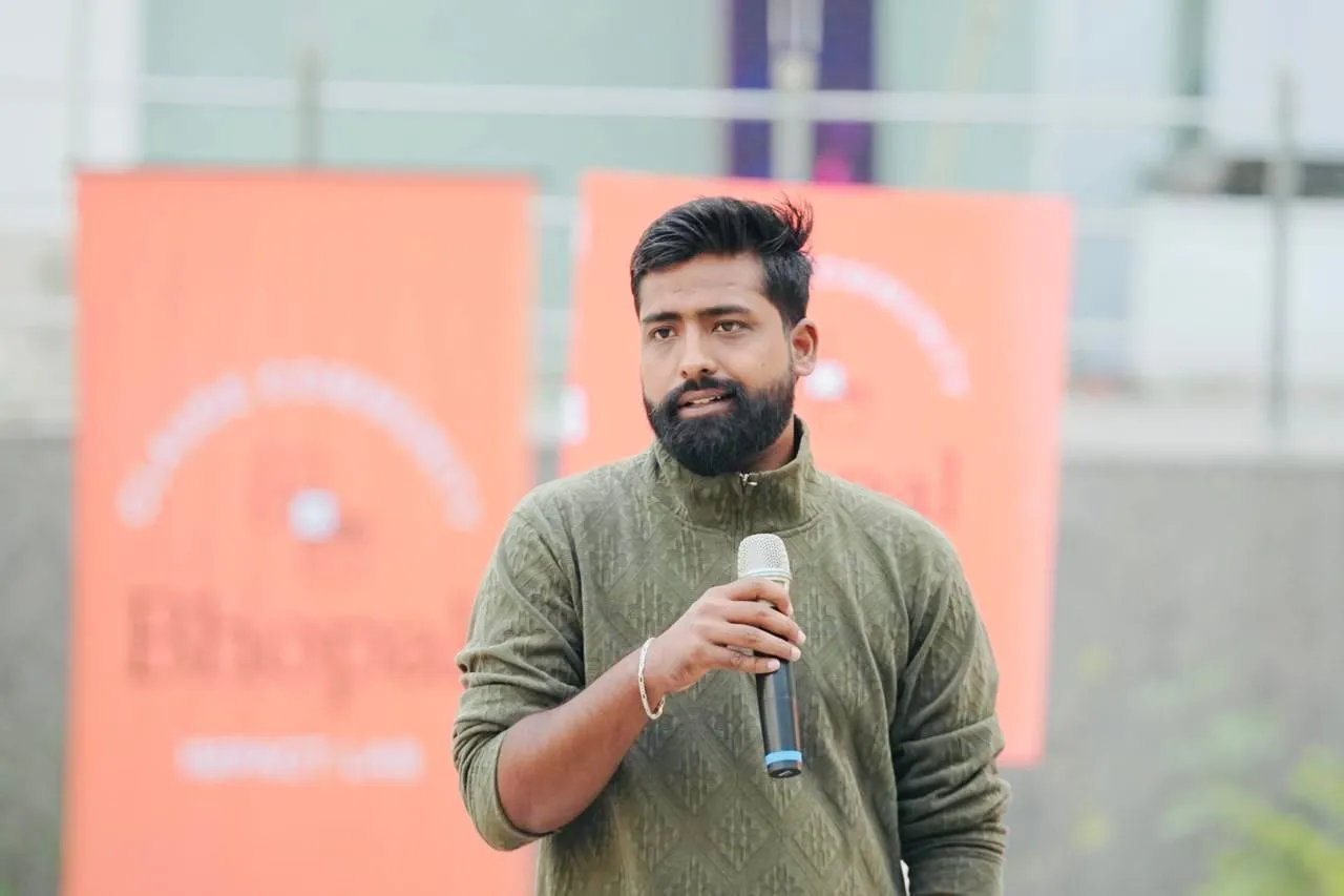 A speaker in a green sweater talking into a microphone in front of orange Claude Community Bhopal banners