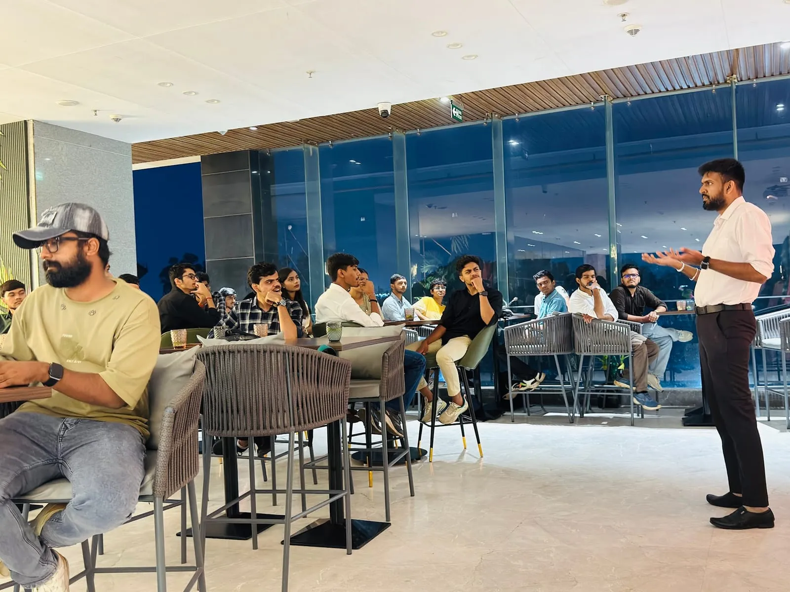 A speaker addressing attendees at lounge tables, with city lights through the glass wall behind them
