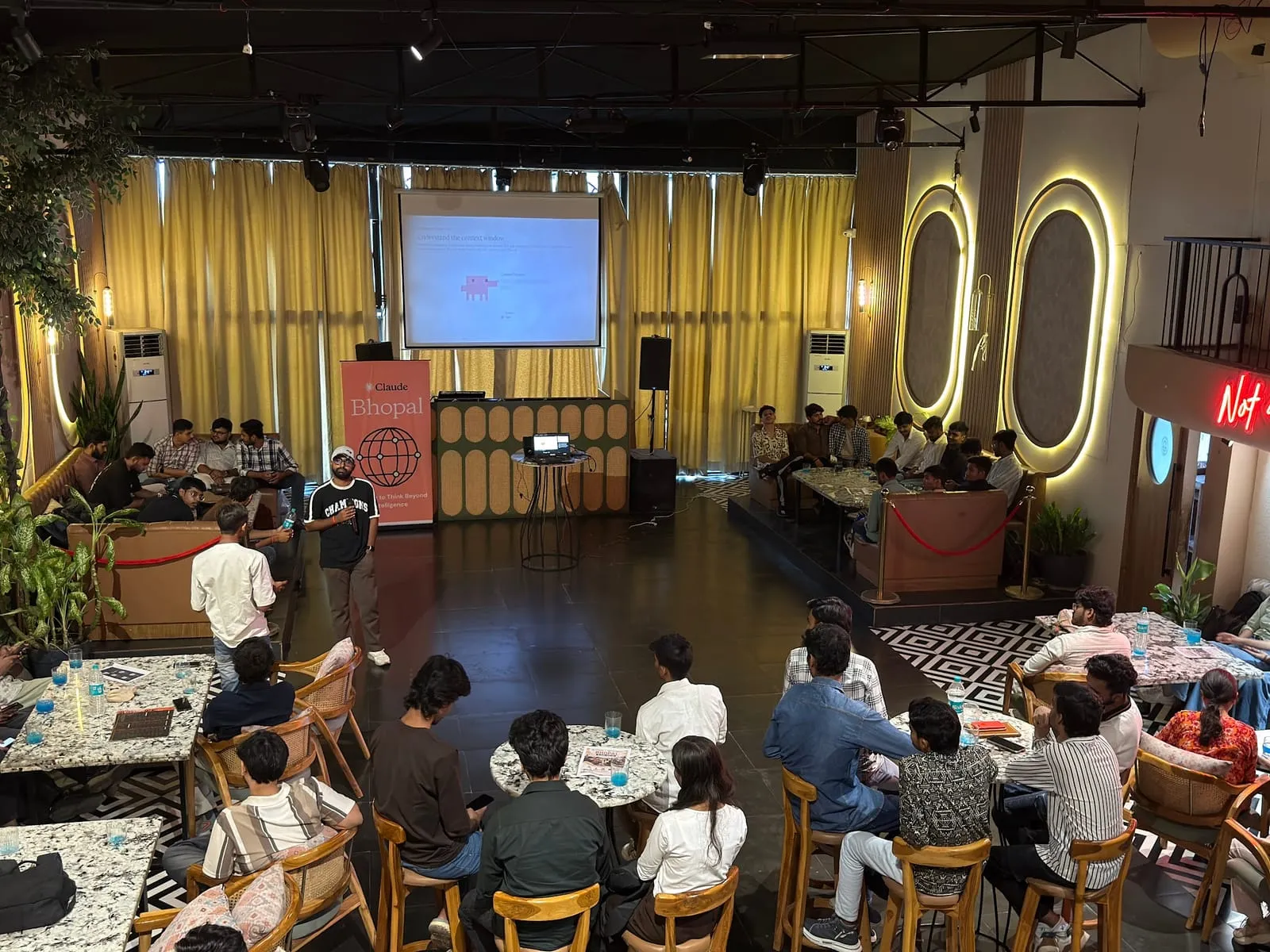 The lounge venue of the Claude Meetup seen from the balcony, attendees at tables facing the host beside the Claude Bhopal banner and the screen