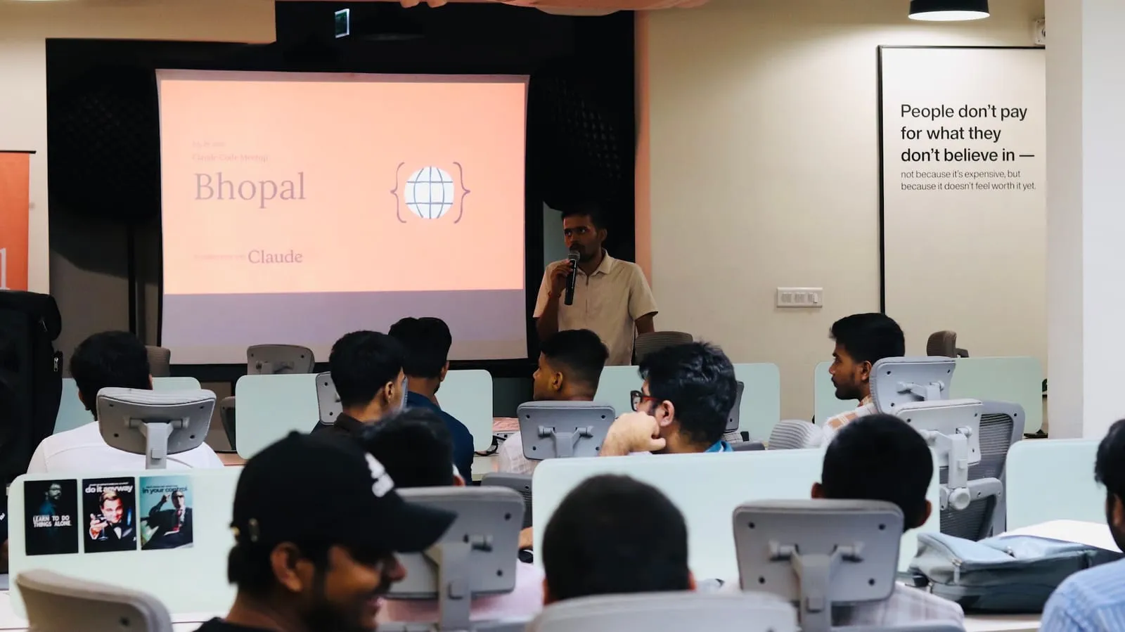 A speaker with a microphone beside the Claude Bhopal title slide, attendees at desks in the foreground