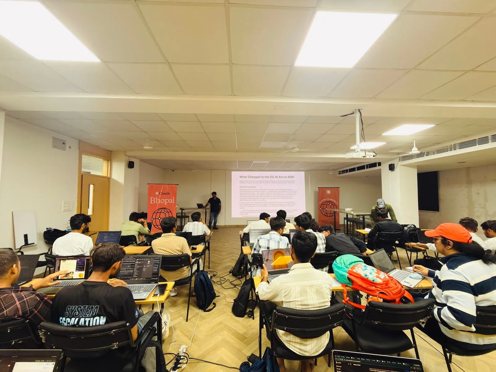 Participants at laptops while a speaker presents a slide at the front of the classroom, between two Claude Bhopal banners