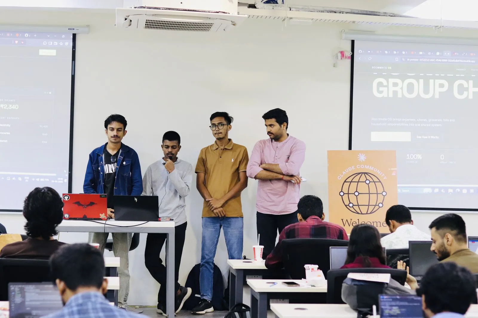 Four participants pitching their project beside the Claude Community standee at the Claude Code Workshop, with the audience at laptops
