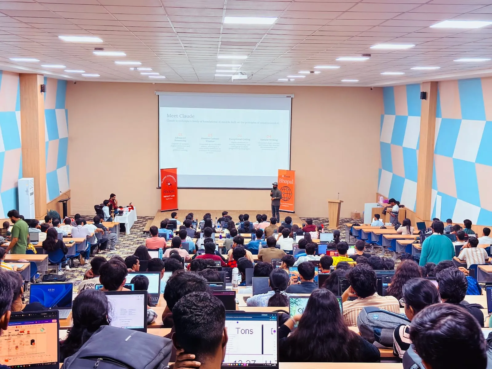 A packed auditorium of students on laptops facing a Meet Claude slide, with a speaker between two Claude Bhopal banners