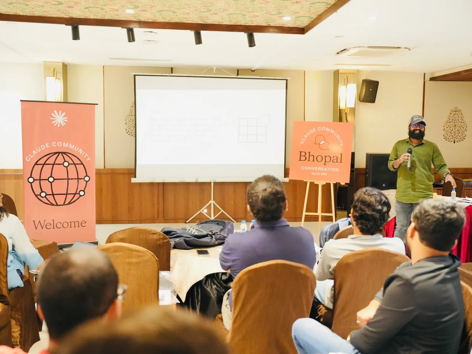The host speaking with a microphone between the Claude Community Welcome banner and the Bhopal Conversation board, seen from behind the audience