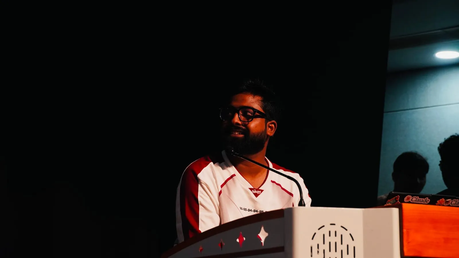 A speaker in a red and white jersey at the lectern in the darkened auditorium