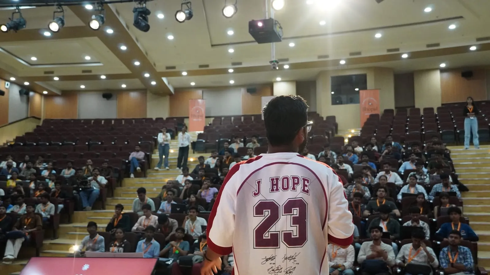 A speaker in a number 23 jersey, seen from behind, facing rows of participants in the auditorium