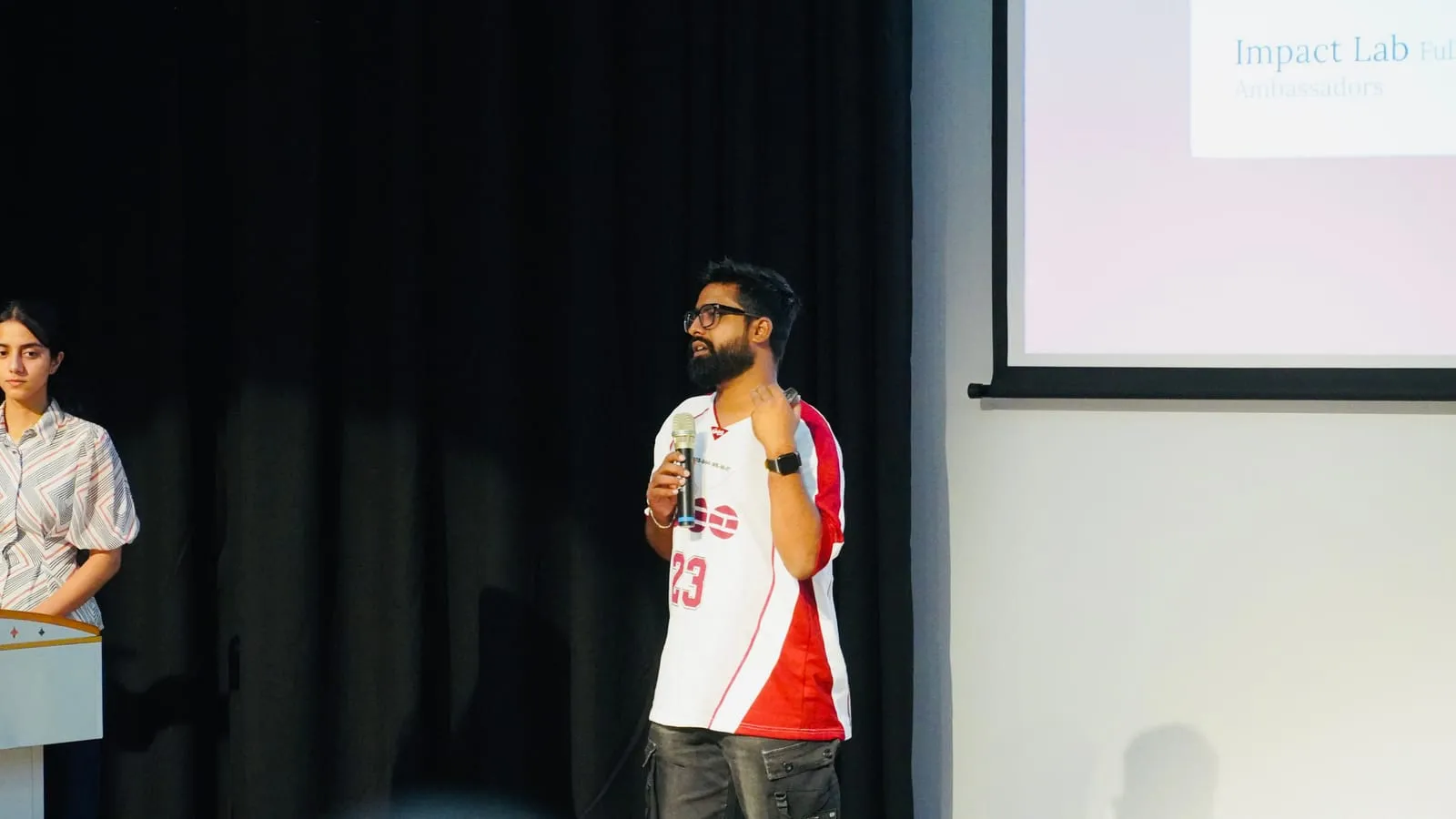 A speaker in a red and white jersey talking into a microphone beside an Impact Lab slide, a co-presenter at the lectern