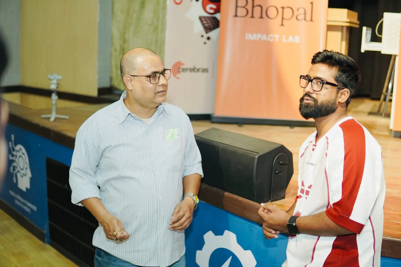 Two people talking beside the stage, a Claude Community Bhopal Impact Lab banner behind them