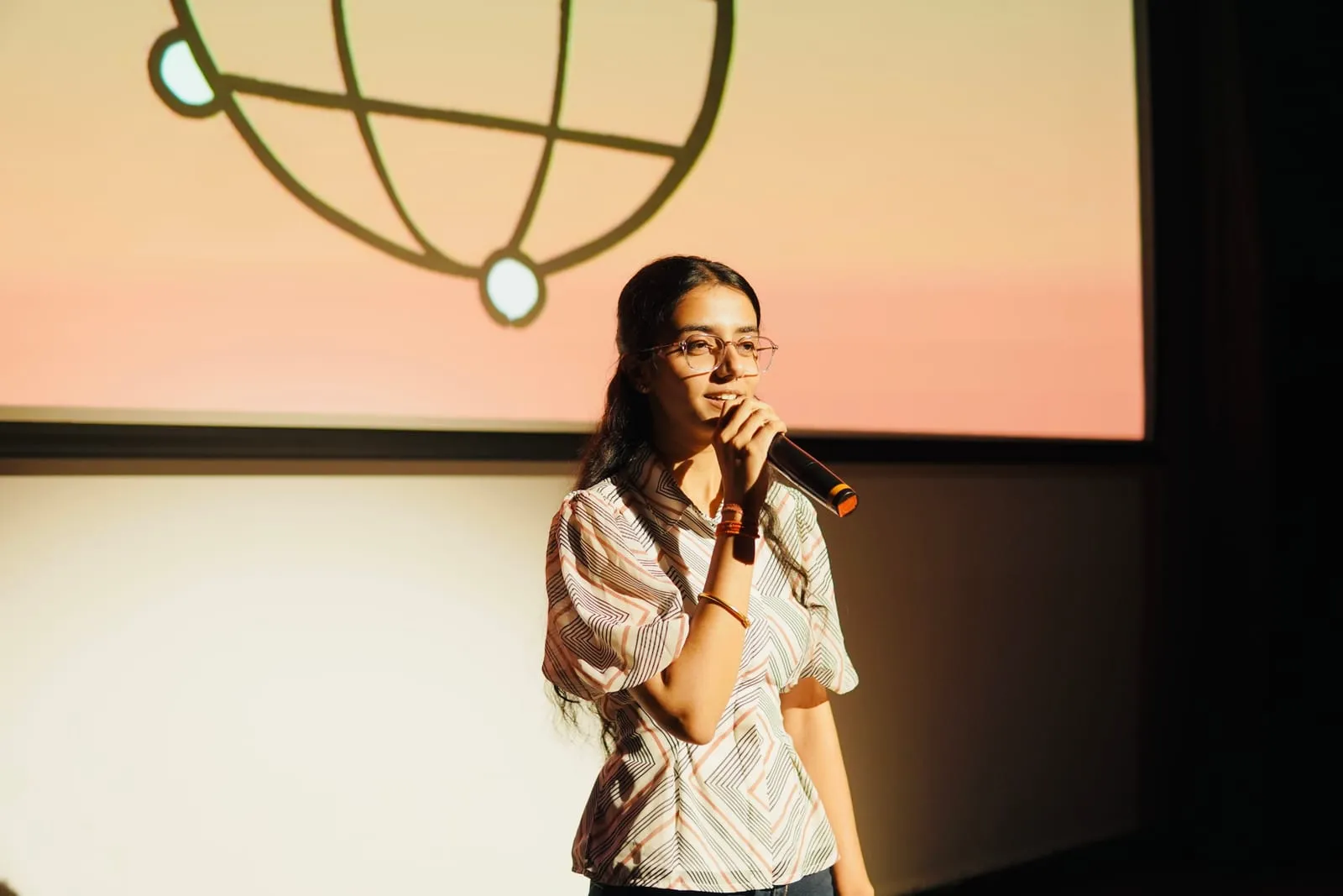 A speaker in a zigzag-patterned blouse talking into a microphone in front of the Claude Community globe on the screen