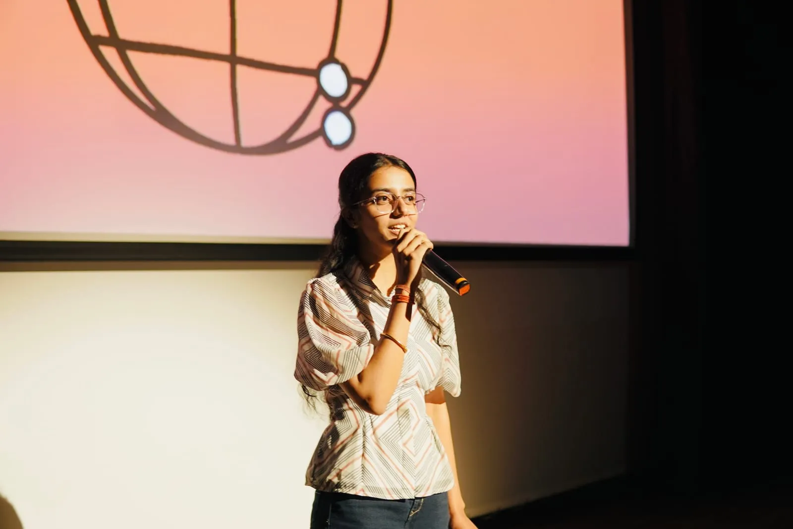 A speaker with a microphone on stage, the globe from the title slide on the screen behind her