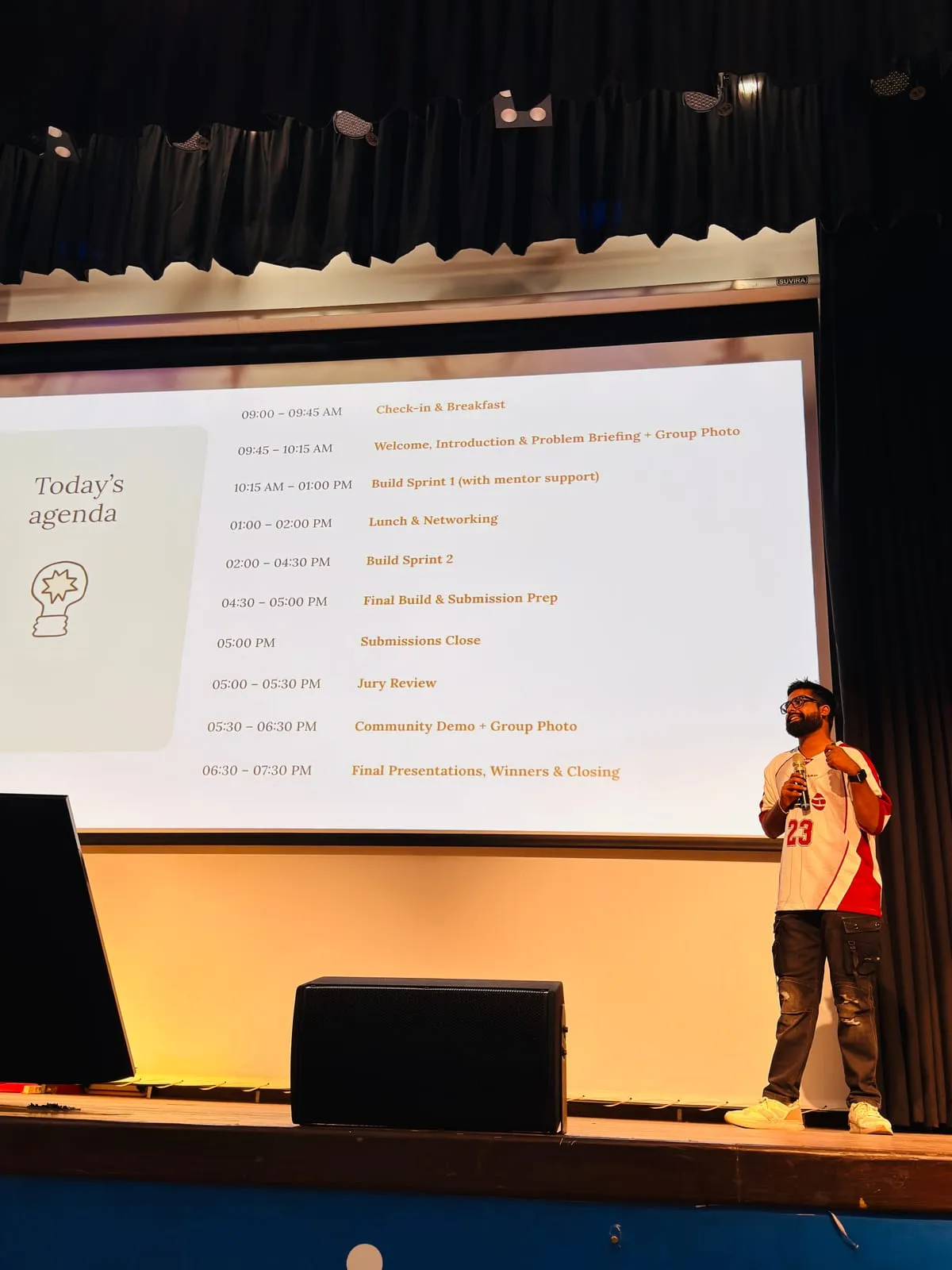 A speaker in a red and white jersey on stage beside the Today’s agenda slide, from check-in and breakfast to the final presentations, winners and closing