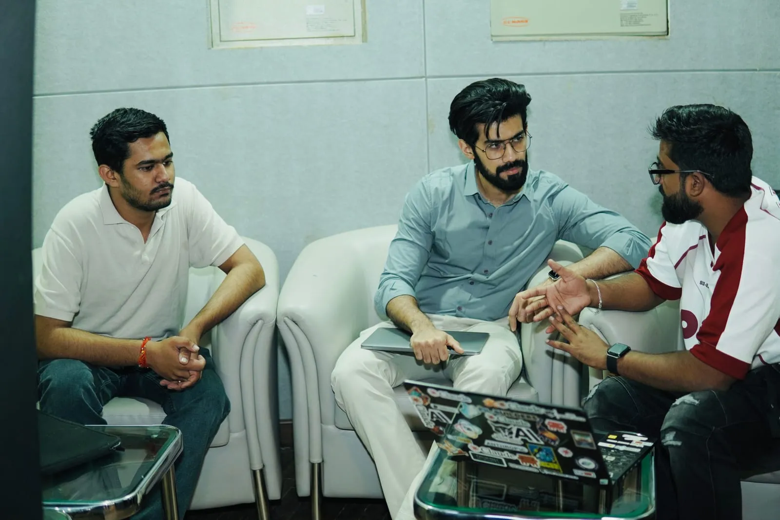 Three people talking in white armchairs, one gesturing over a laptop covered in stickers